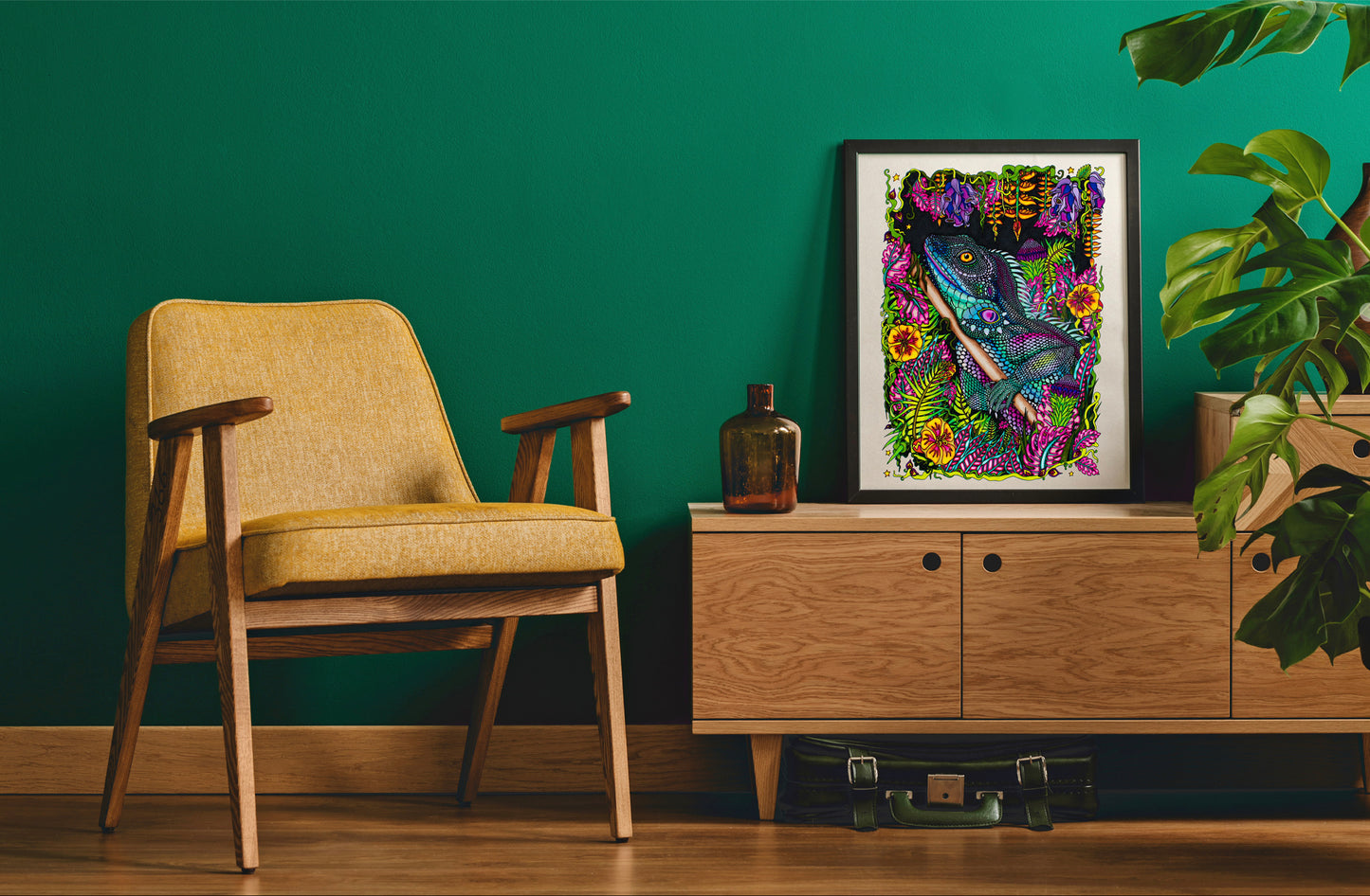 Blue Iguana in the jungle in a black frame in a green room. There is a bottle on the table and an old suitcase on the floor under the sideboard. There is a yellow chair and a cheese plant.